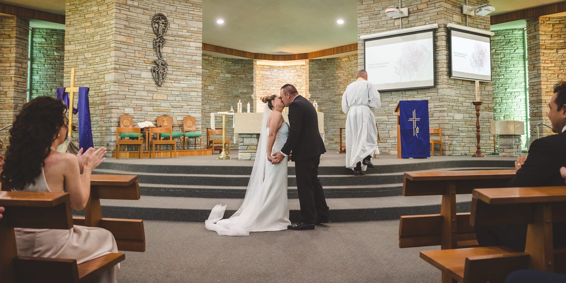 Bride and groom kiss at an altar in a stone chapel. Guests seated, clapping. Blue cloth with cross in background. Joyful atmosphere.
