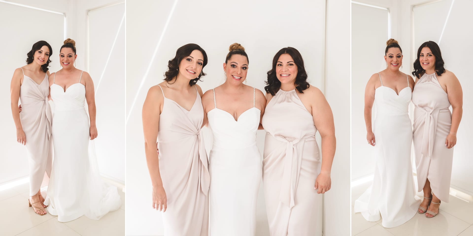 Three women in elegant gowns stand together smiling in a bright room with white walls. The central woman wears a bridal dress.