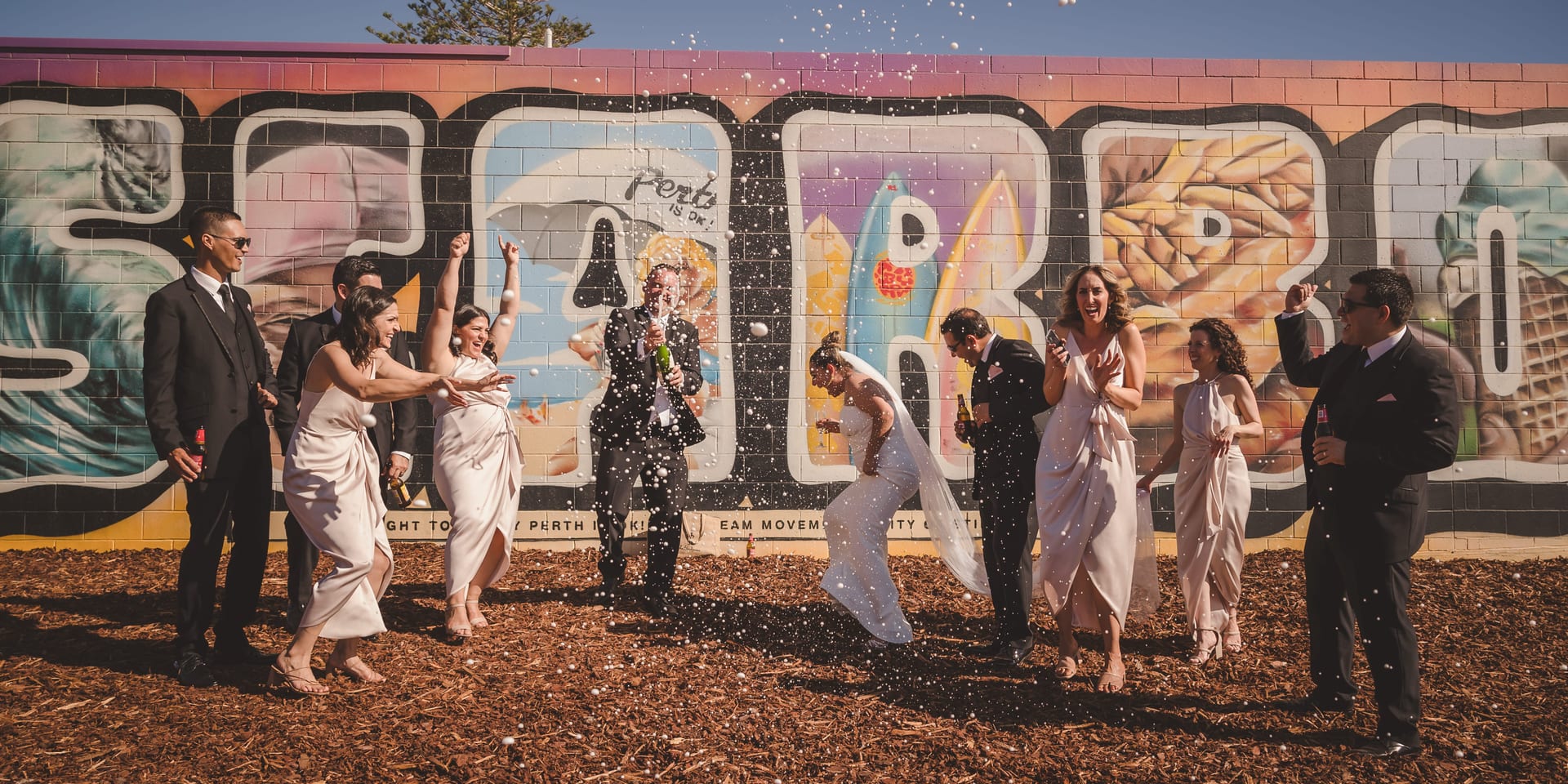 Wedding party celebrating with champagne in front of colorful graffiti wall. People in formal attire, joyful mood, sunny day.