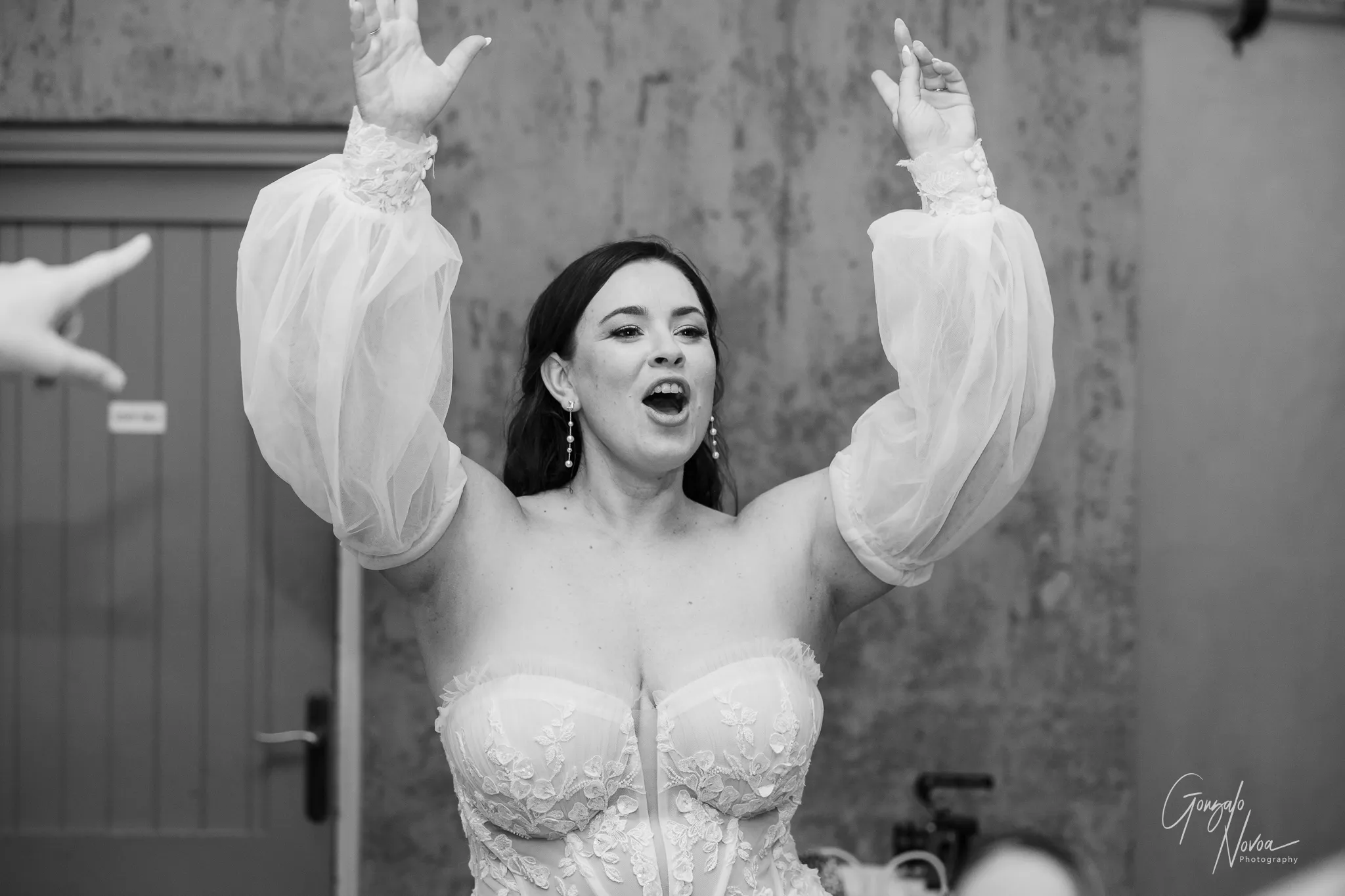 The bride, wearing a lace off-shoulder wedding dress, lifts her arms high while singing joyfully during the reception. Her expression is full of energy and excitement.