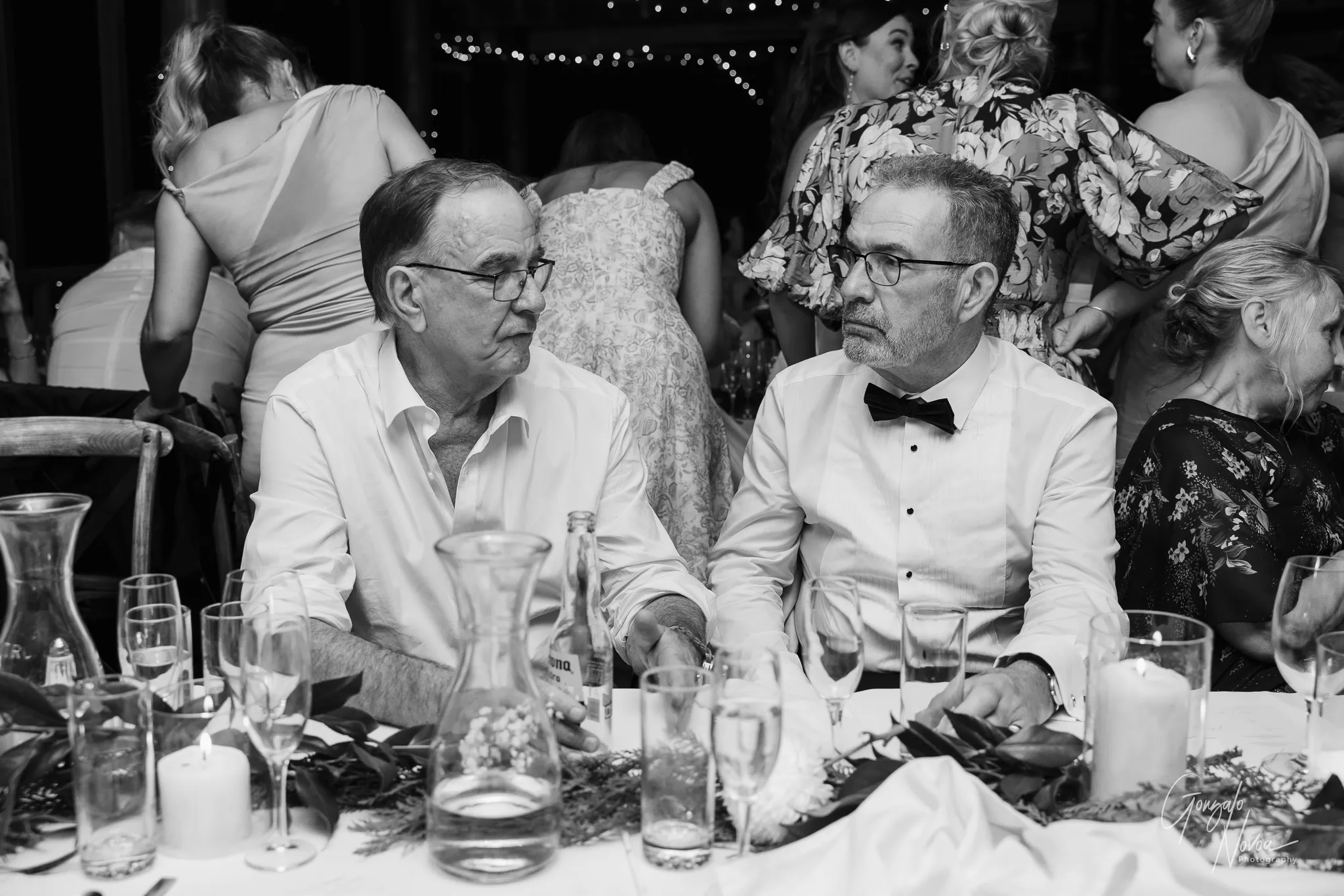 The father of the bride and the father of the groom sit together at the wedding reception, sharing a conversation. One wears a white shirt with rolled-up sleeves and glasses, holding a beer, while the other wears a formal white shirt with a black bow tie and glasses. They are surrounded by an elegantly decorated table with candles, glasses, and greenery, while guests mingle in the background under fairy lights.