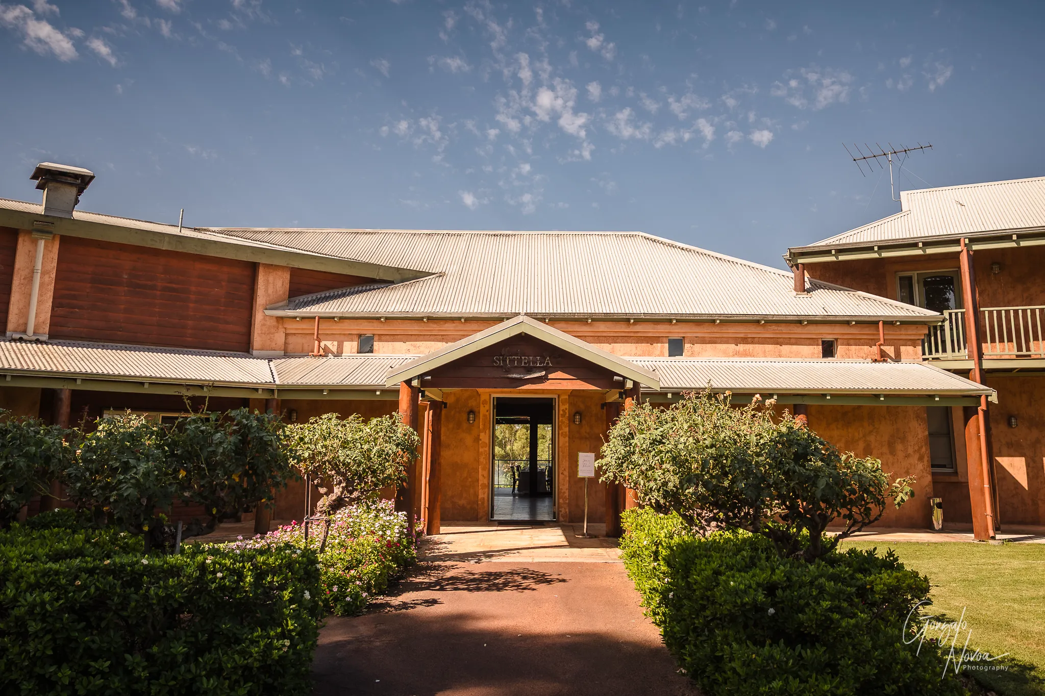 The front entrance of Sittella Winery, featuring rustic terracotta-coloured walls, a corrugated metal roof, and lush gardens lining the pathway under a clear blue sky.