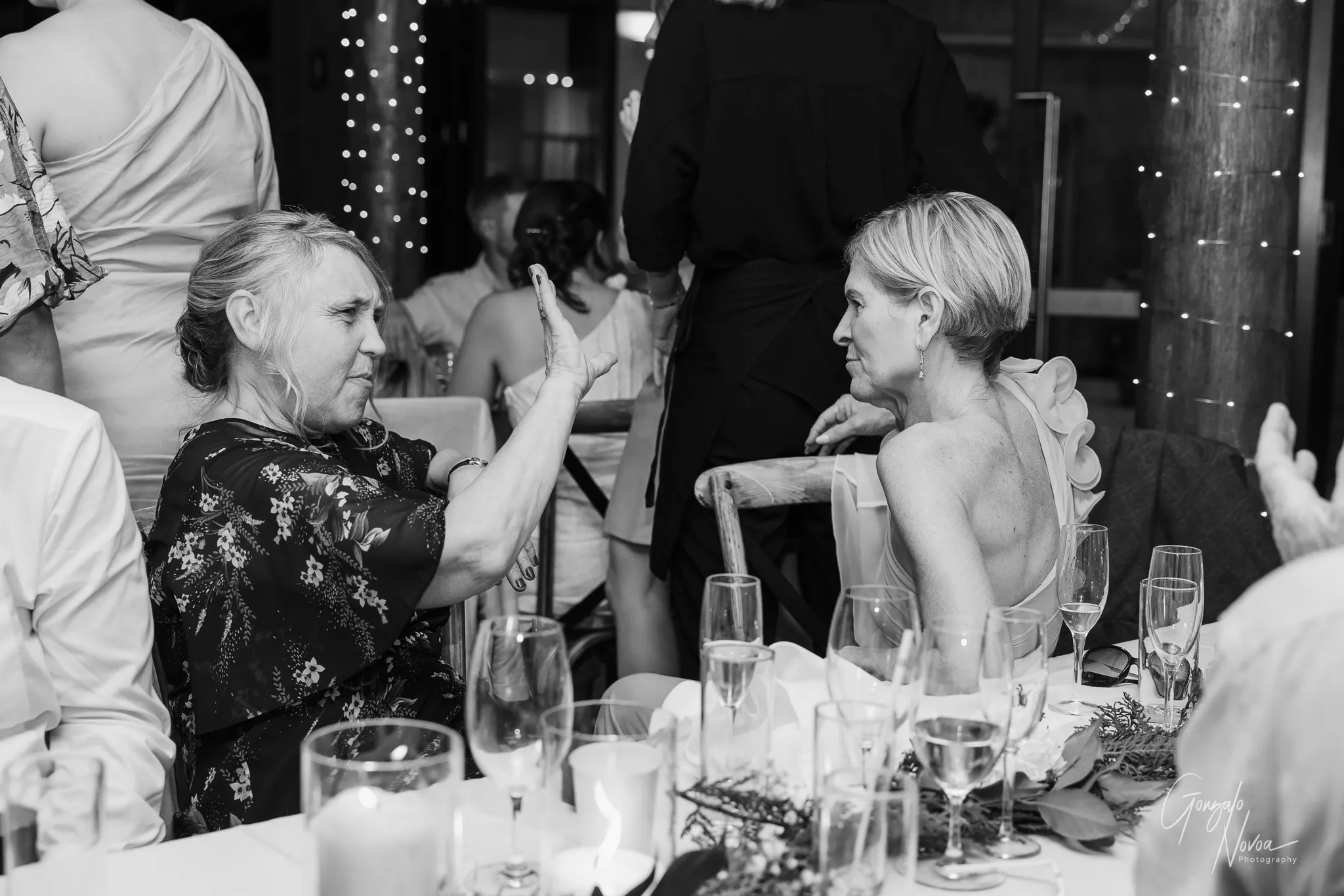 The mother of the bride and the mother of the groom share an animated conversation at the wedding reception. One wears a floral-patterned dress and gestures expressively with her hand, while the other, in a sleeveless gown with floral details on the shoulder, listens intently. They are seated at a decorated table with champagne flutes, candles, and greenery, surrounded by guests and fairy lights.