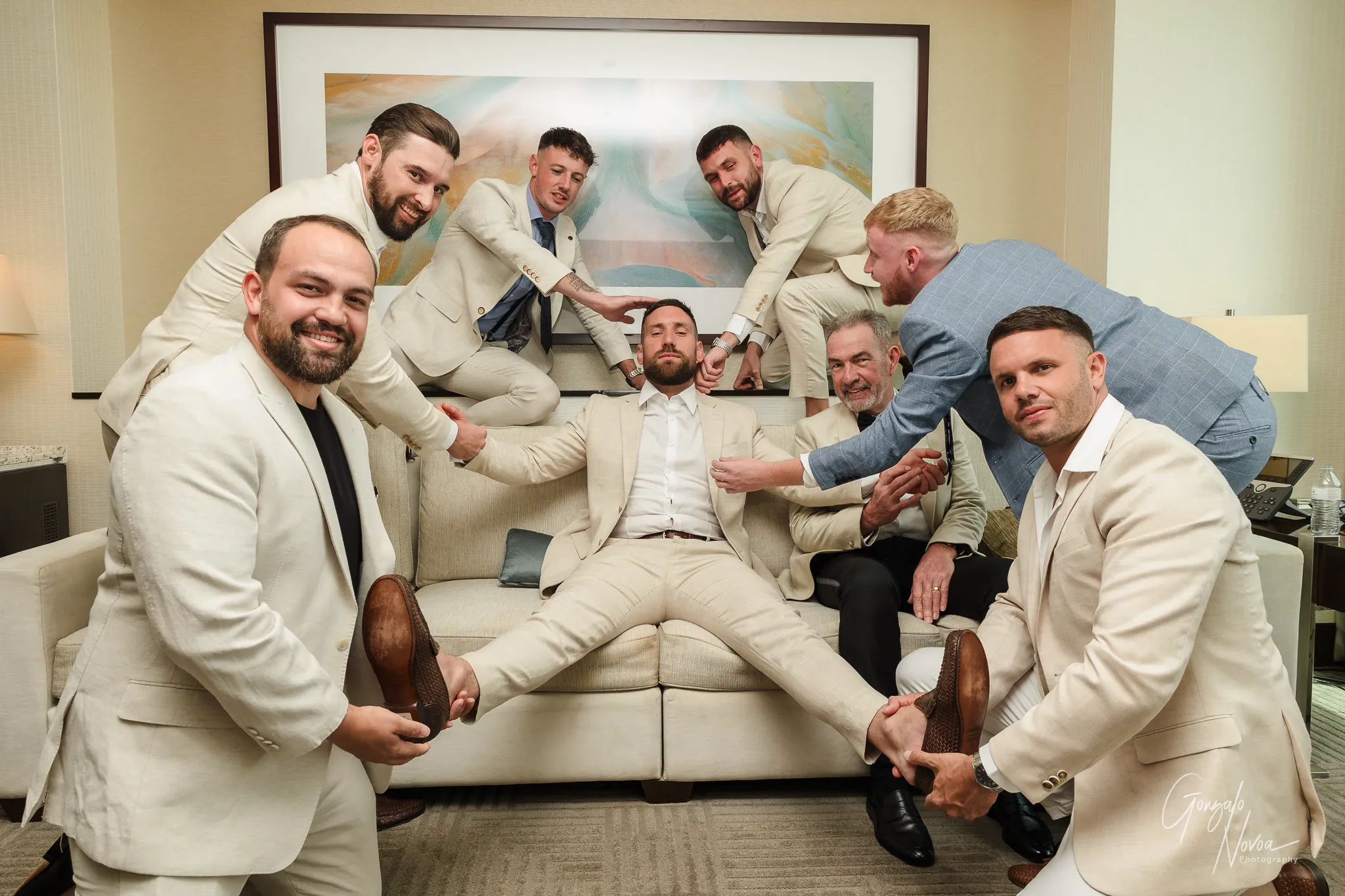 Playful group photo of James lounging on a hotel couch while his groomsmen, friends and dad surround him, adjusting his suit, shoes and tie in a fun, exaggerated way. Everyone is laughing and smiling in a relaxed pre-wedding moment at Crown Towers Perth.