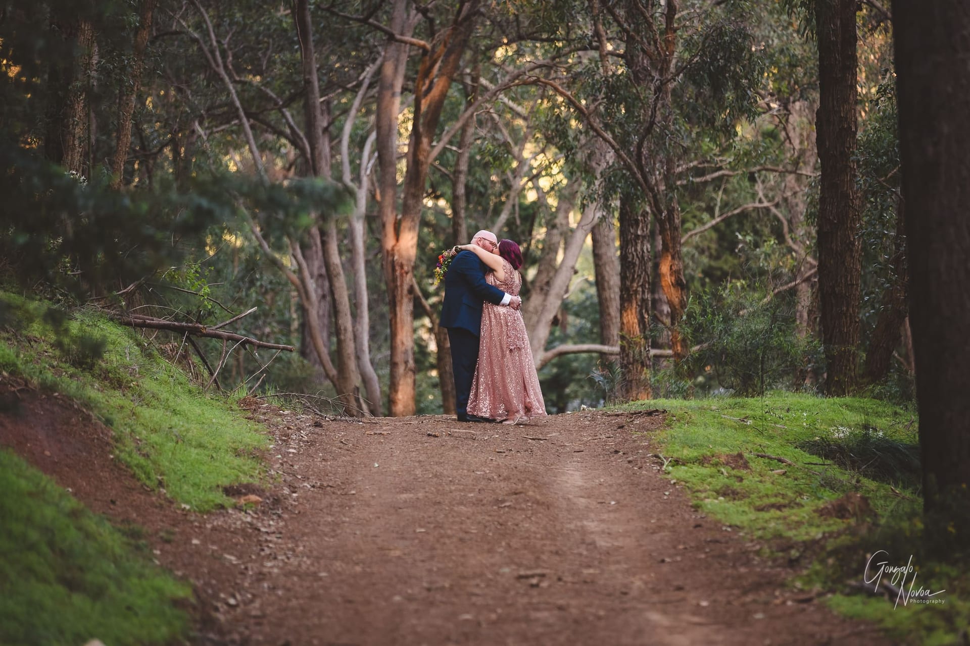 Bride and groom, embraces on a dirt path in the woods, surrounded by tall trees and a serene atmosphere.