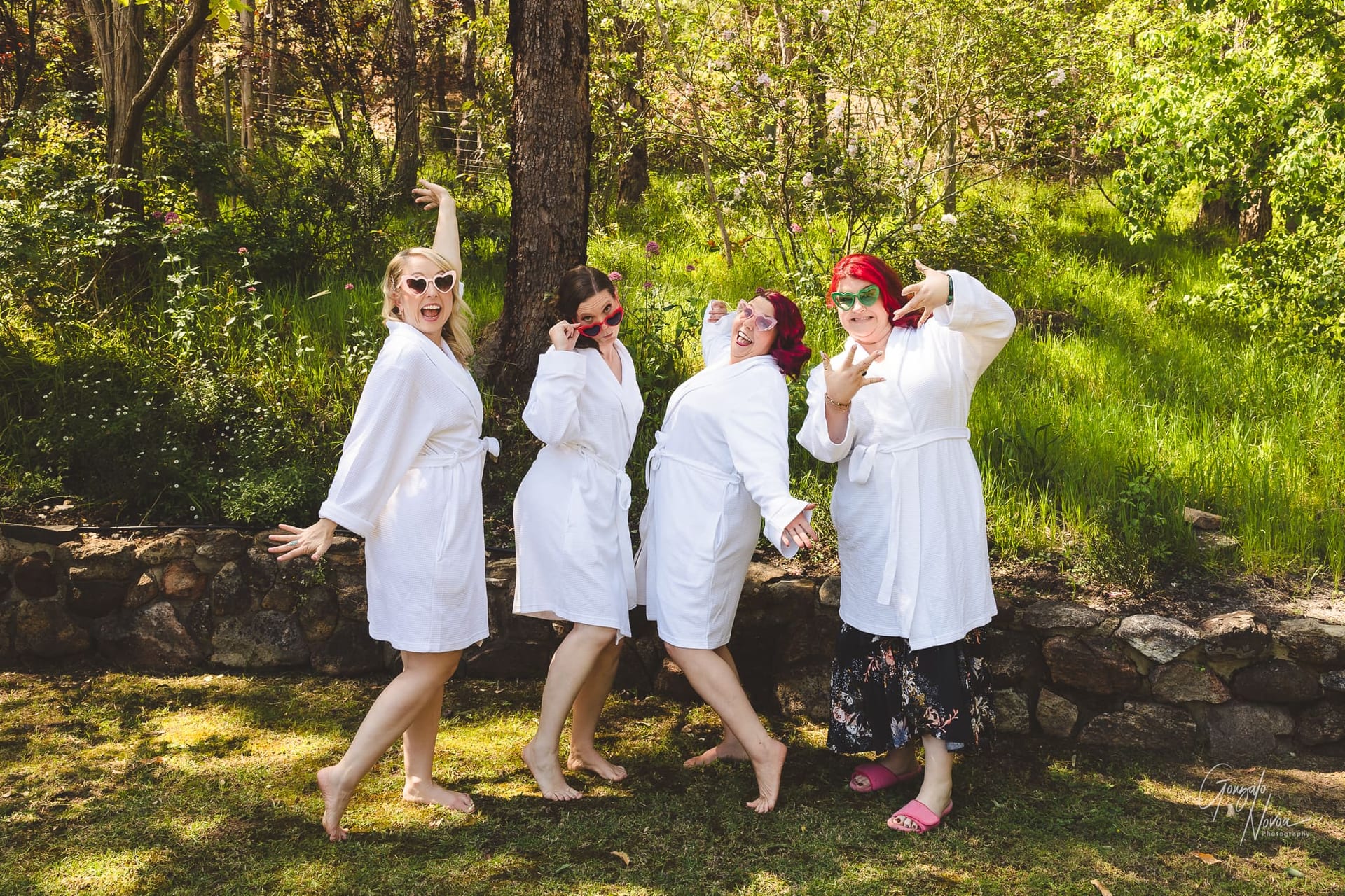 Photo of four bridesmaids in white robes posing cheekily with big smiles, and fun style sunglasses