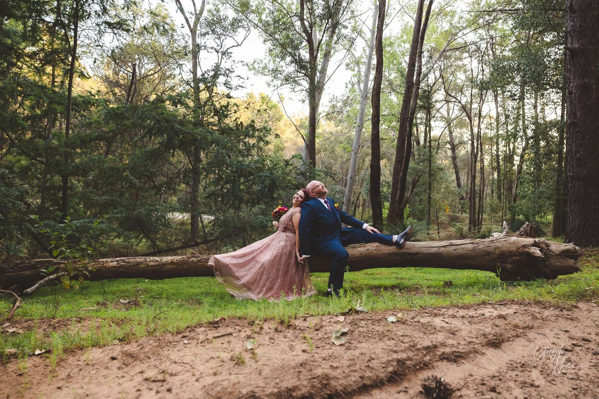 Serene moment in the woods. The bride and groom rest against each other's back while sitting over a fallen tree trunk