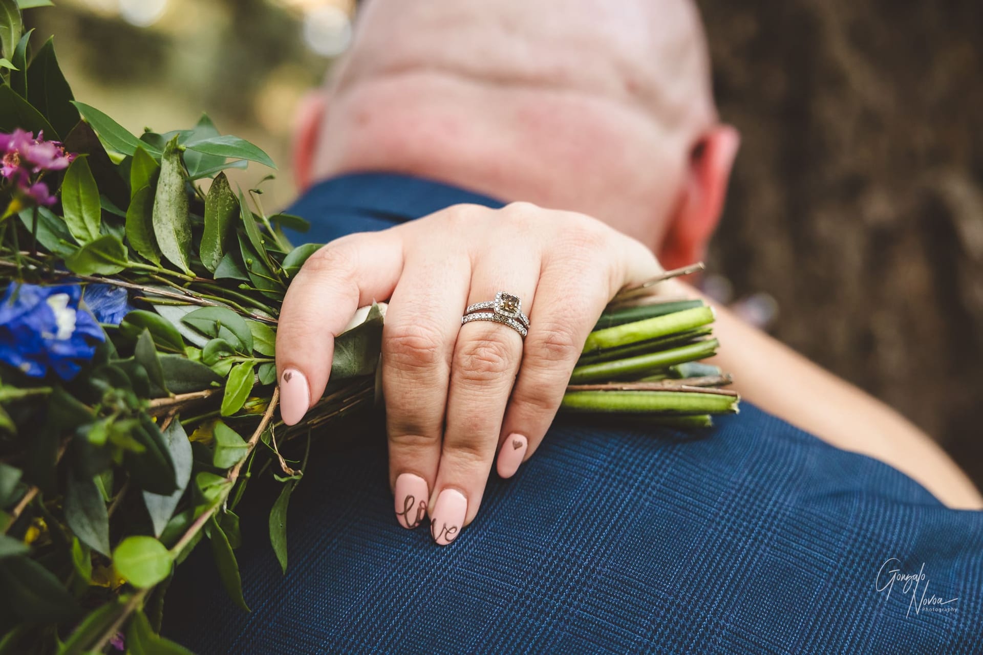 Wedding ring and love nails! Photo from behind of the bride and groom where the bride's hand can be seen hugging the groom, with a close up of her hand and ring in sight, the nail polish reads love across the middle and ring finger