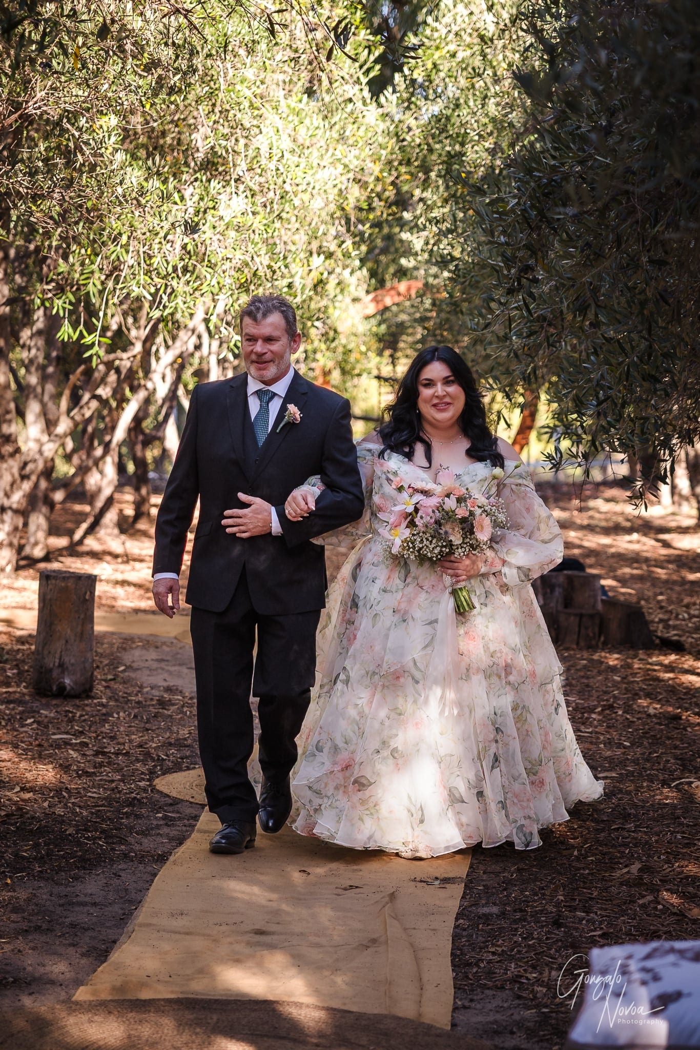 Bride in floral dress walks arm-in-arm with her father in a suit down a wooded path. She holds a bouquet. Sunlight filters through leaves.
