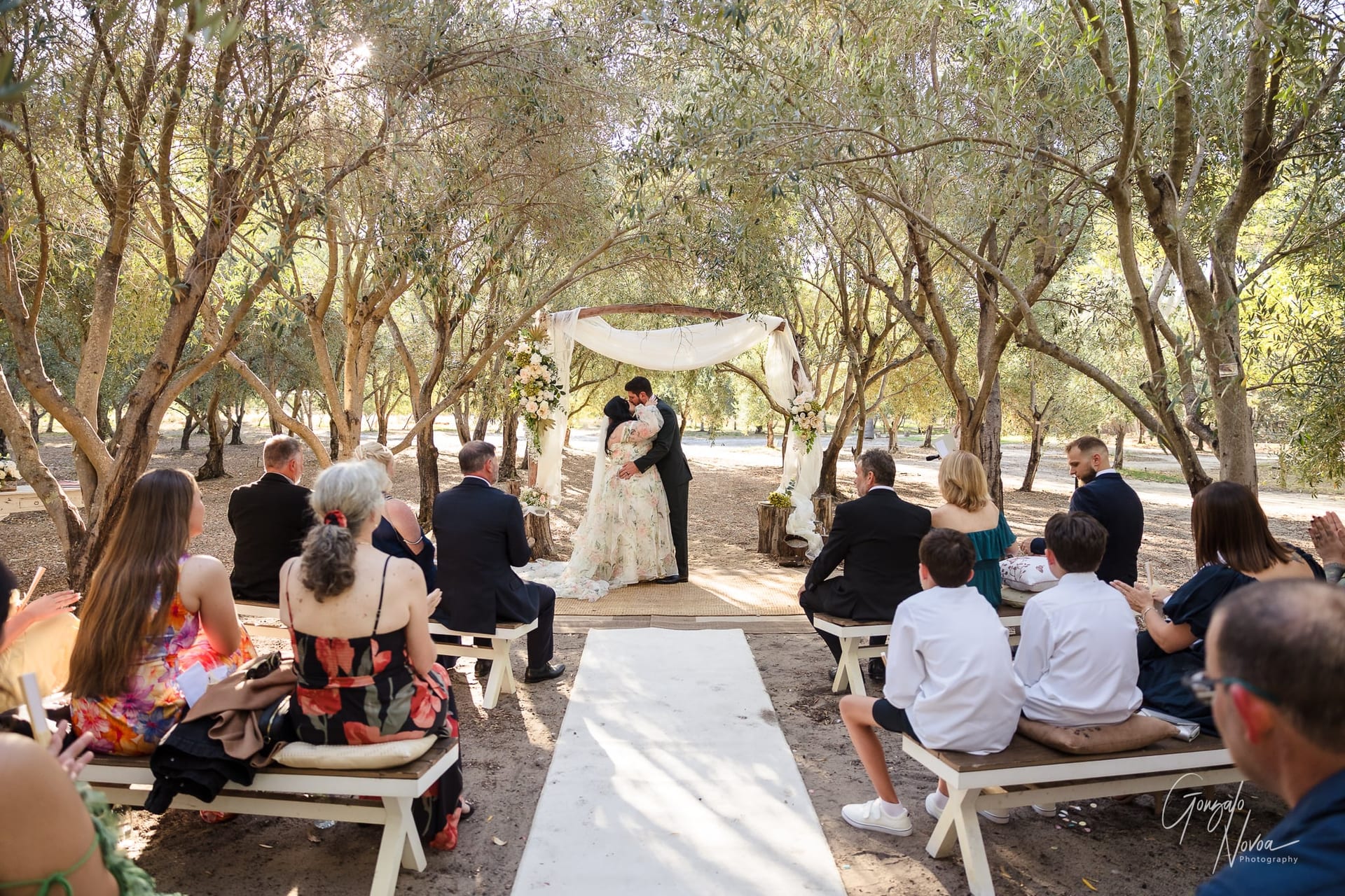 A couple embraces under a floral arch in a sunlit outdoor wedding. Guests sit on benches among trees, clapping. Text: Gonzalo Novoa Photography.