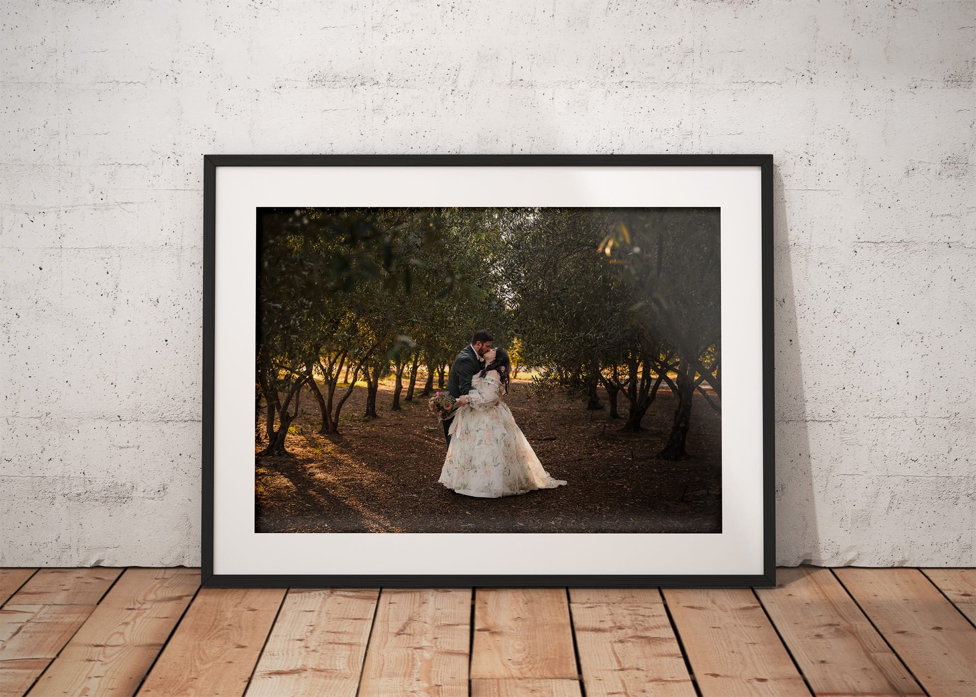 Framed photo of a bride and groom embracing in an olive grove, bathed in soft golden-hour light, displayed against a white wall and wooden floor.