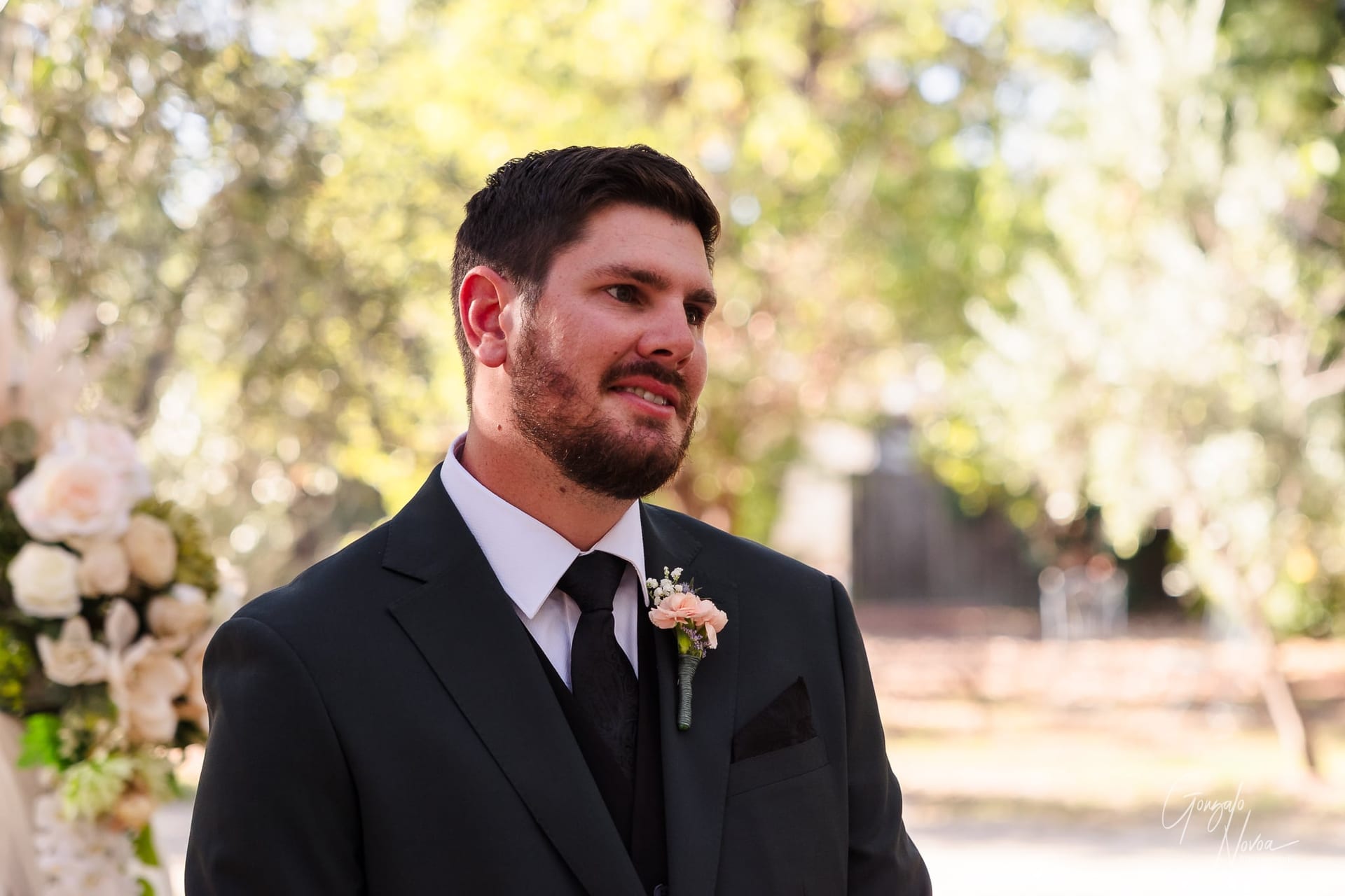 Groom in a suit with a floral boutonniere, smiling at an outdoor event. Blurred trees in the background, soft natural light.