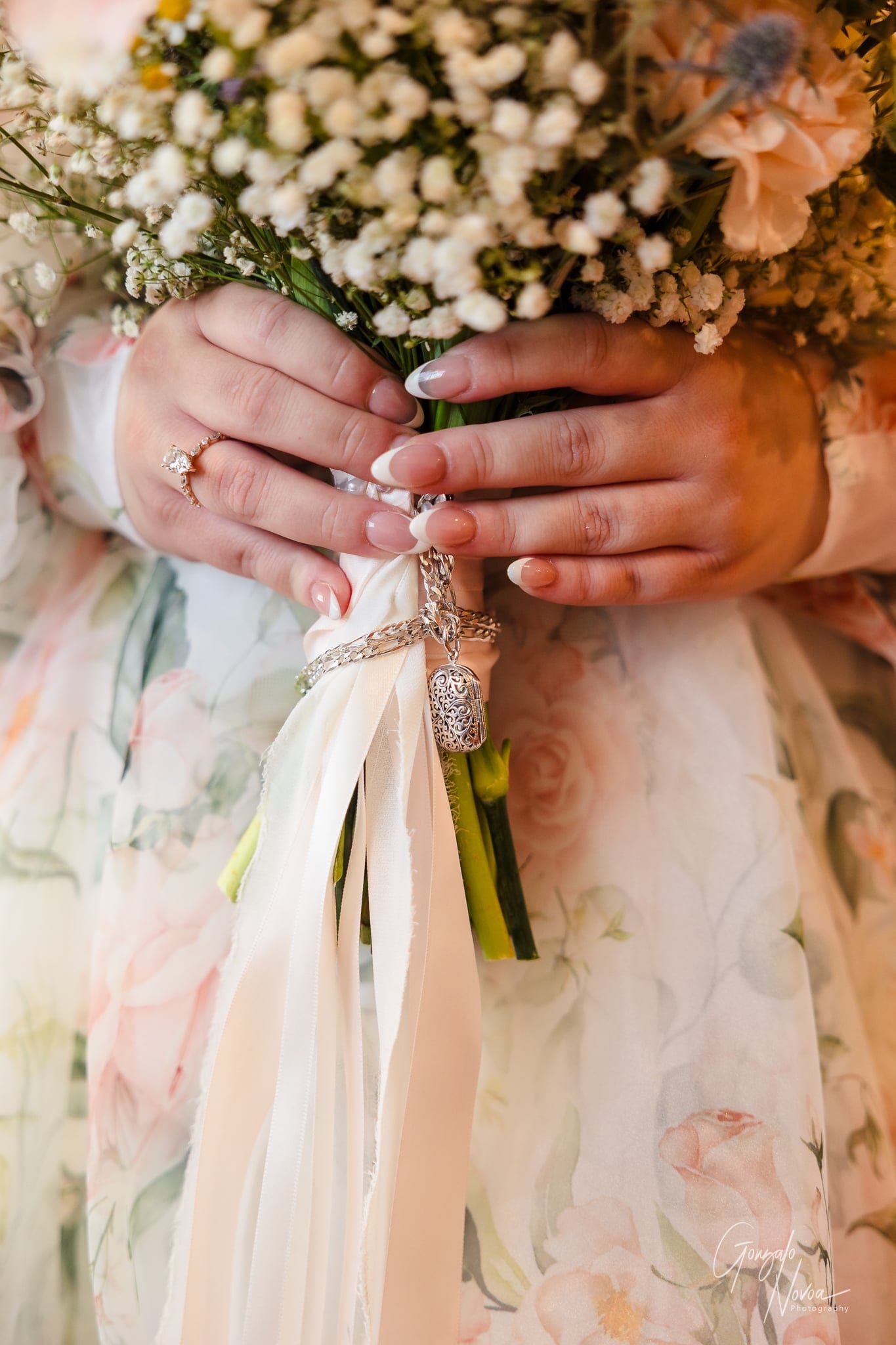Hands with a diamond ring holding a bouquet of white flowers with ribbon, against a floral-patterned dress. Warm, delicate mood.