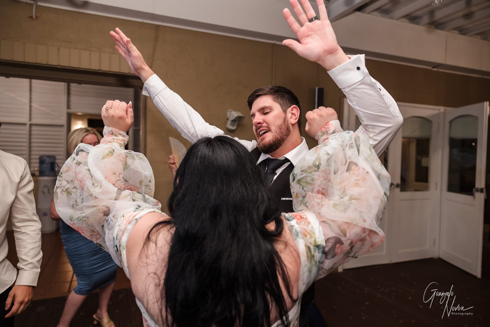 People joyfully dance with raised arms at an indoor event. The woman wears a floral dress, and the man is in a suit. Background is neutral.