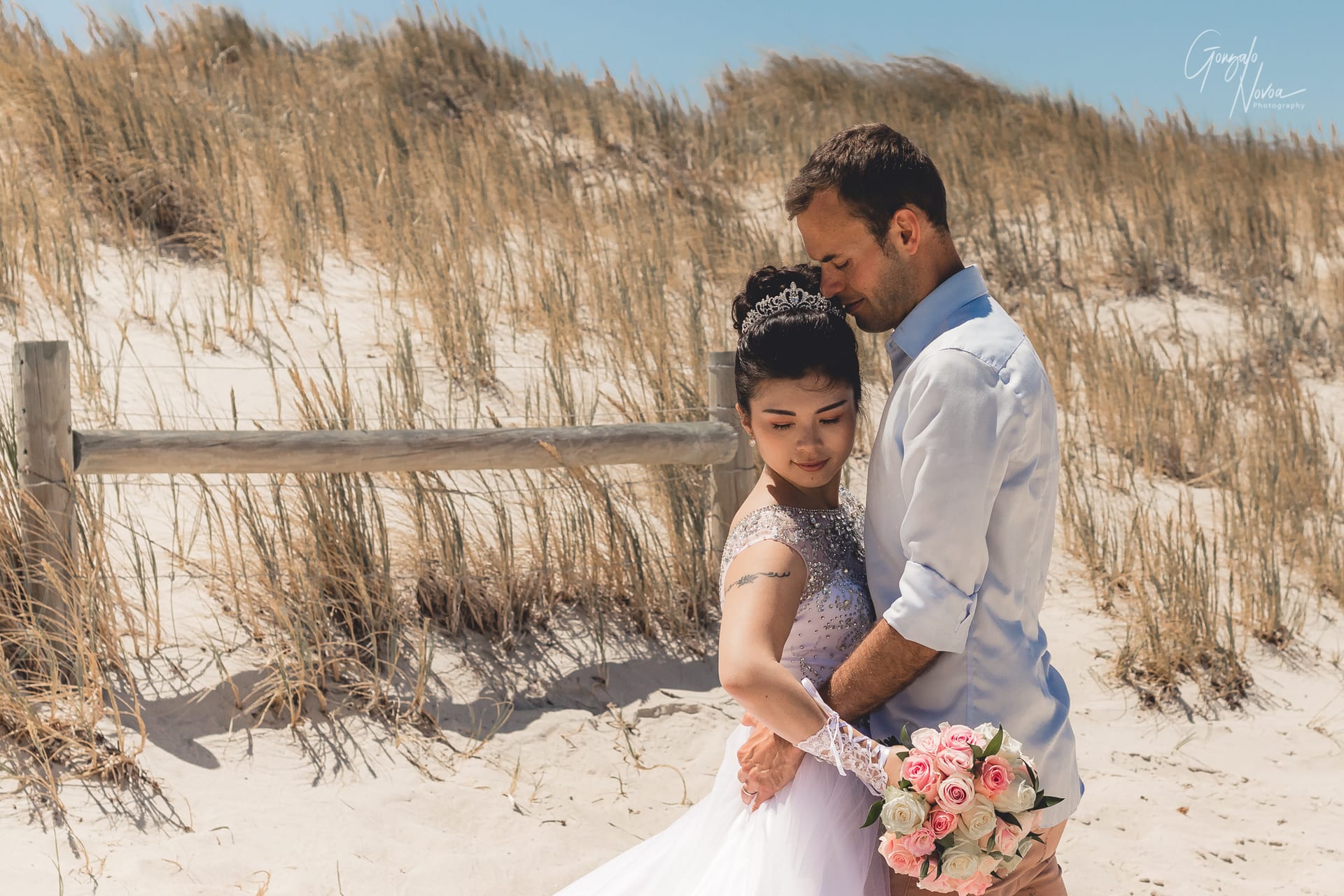 Bride and groom share a quiet, intimate moment during their First Look on a sandy beach, with the bride holding a bouquet of pink and white roses.